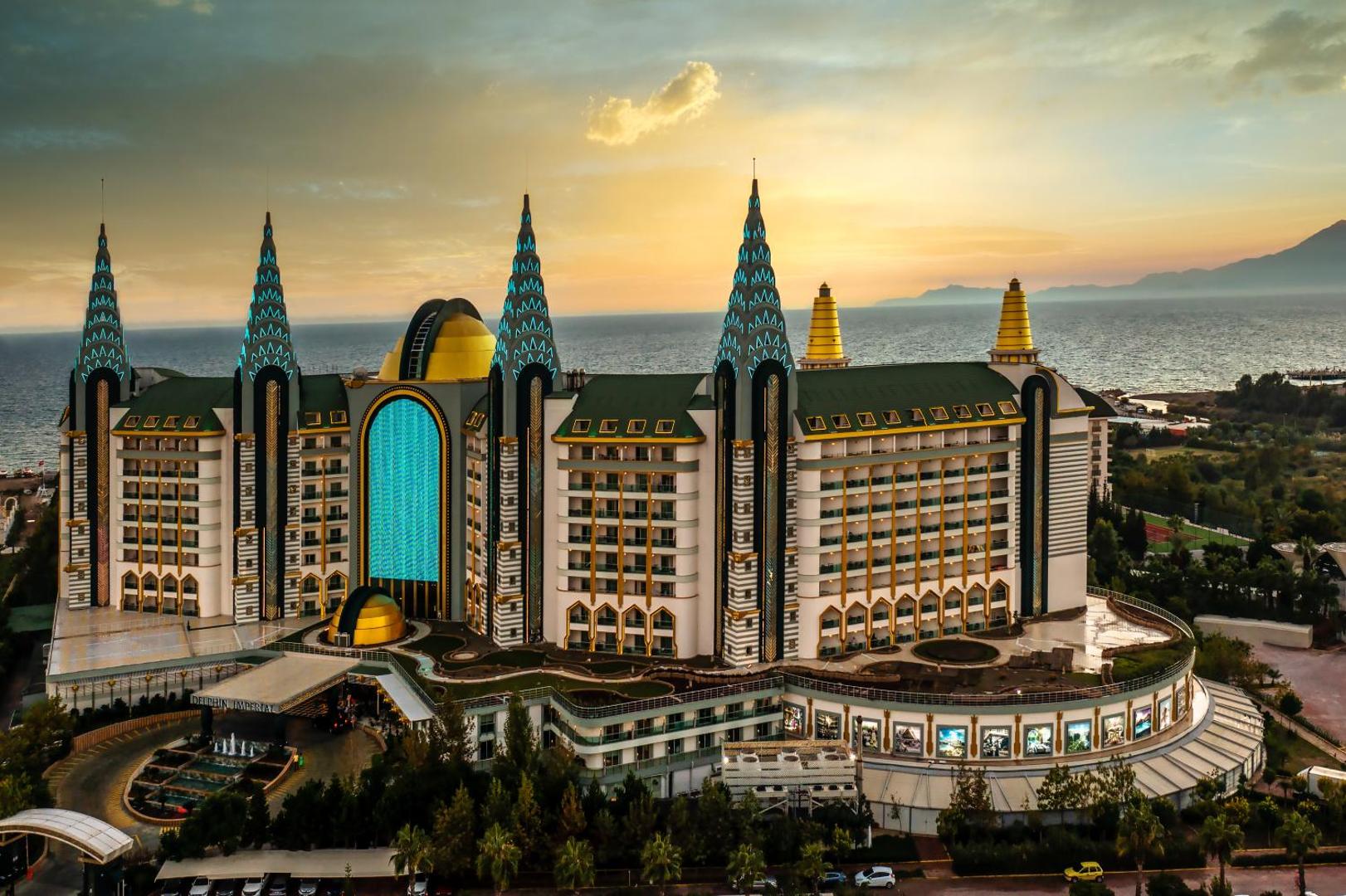 Delphin Imperial Hotel fasāde ar unikāliem torņiem zelta stundā — skats uz viesnīcas korpusiem un labiekārtoto teritoriju