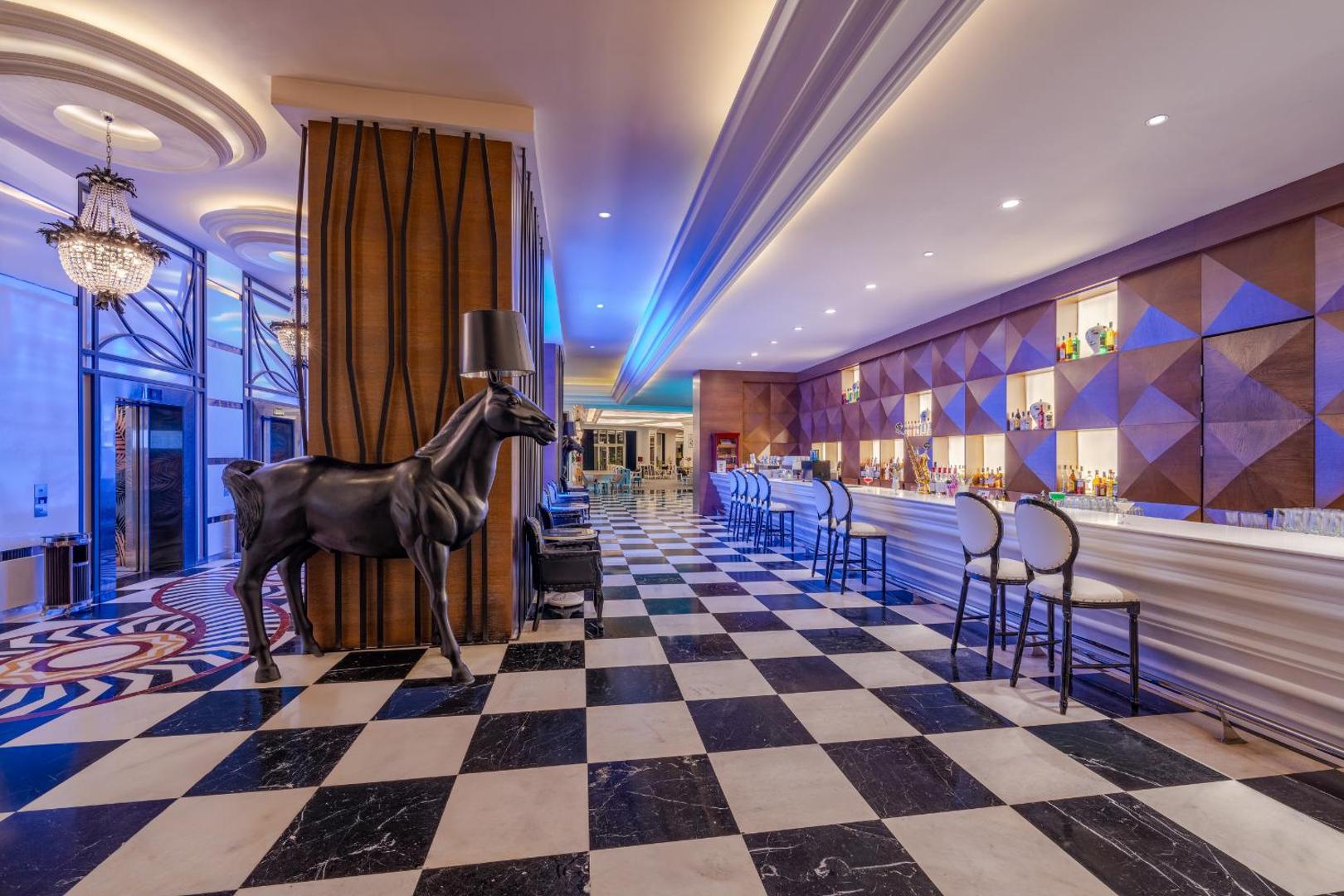 Delphin Imperial Hotel stilīgais bārs ar melnbaltu rūtainu grīdu, modernu leti un dekoratīvu zirga skulptūru