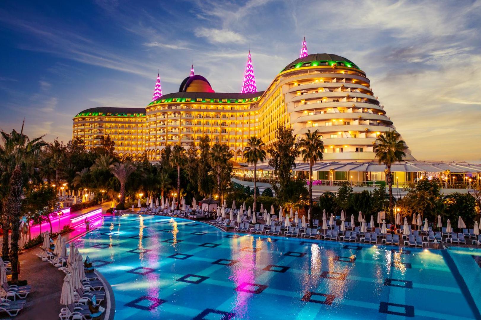 Delphin Imperial Hotel iespaidīgā fasāde un galvenais baseins vakarā — izgaismota arhitektūra, palmas un atpūtas zona pie ūdens Larā