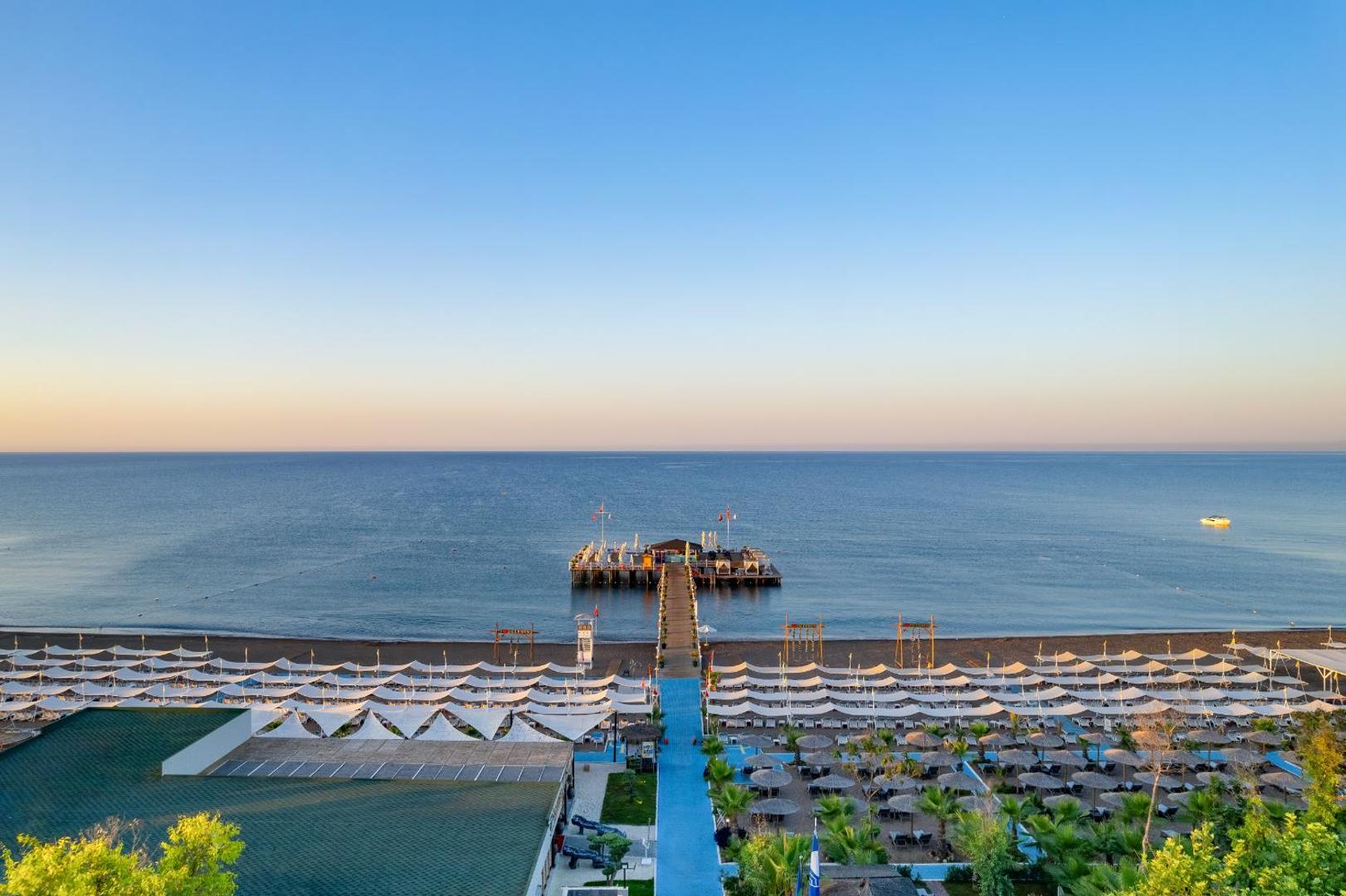 Delphin Imperial Hotel skats no augšas uz privāto pludmali, koka piestātni un Vidusjūru Antaljas piekrastē saulrieta laikā