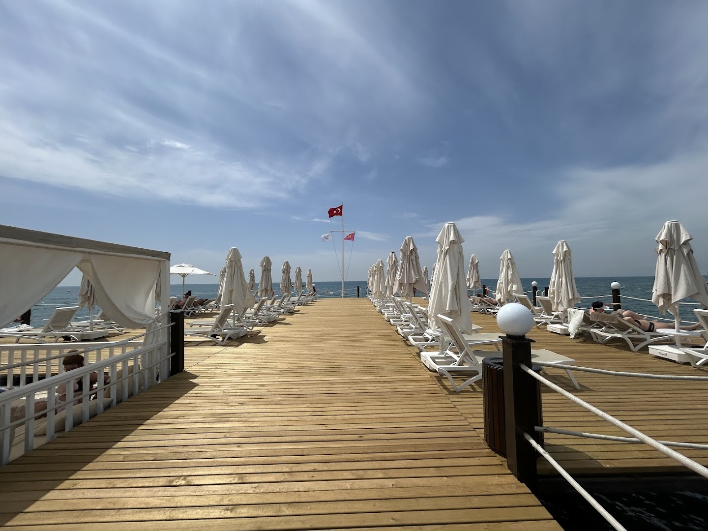 Aska Lara Resort & Spa koka mols ar sauļošanās krēsliem un saulessargiem virs dzidrā jūras ūdens Antaljas reģionā