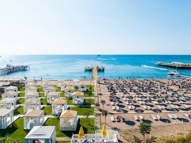 Aska Lara Resort & Spa plašā smilšu pludmale un zāliena zona ar baltiem bungalo un saulessargiem Antaljas piekrastē, Turcijā