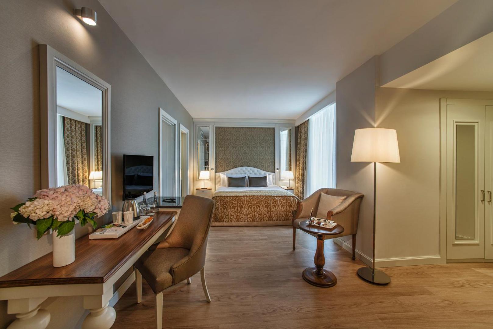 Aska Lara Resort & Spa viesnīcas numura atpūtas zona ar rakstāmgaldu, spoguli un mīksto krēslu siltos toņos.