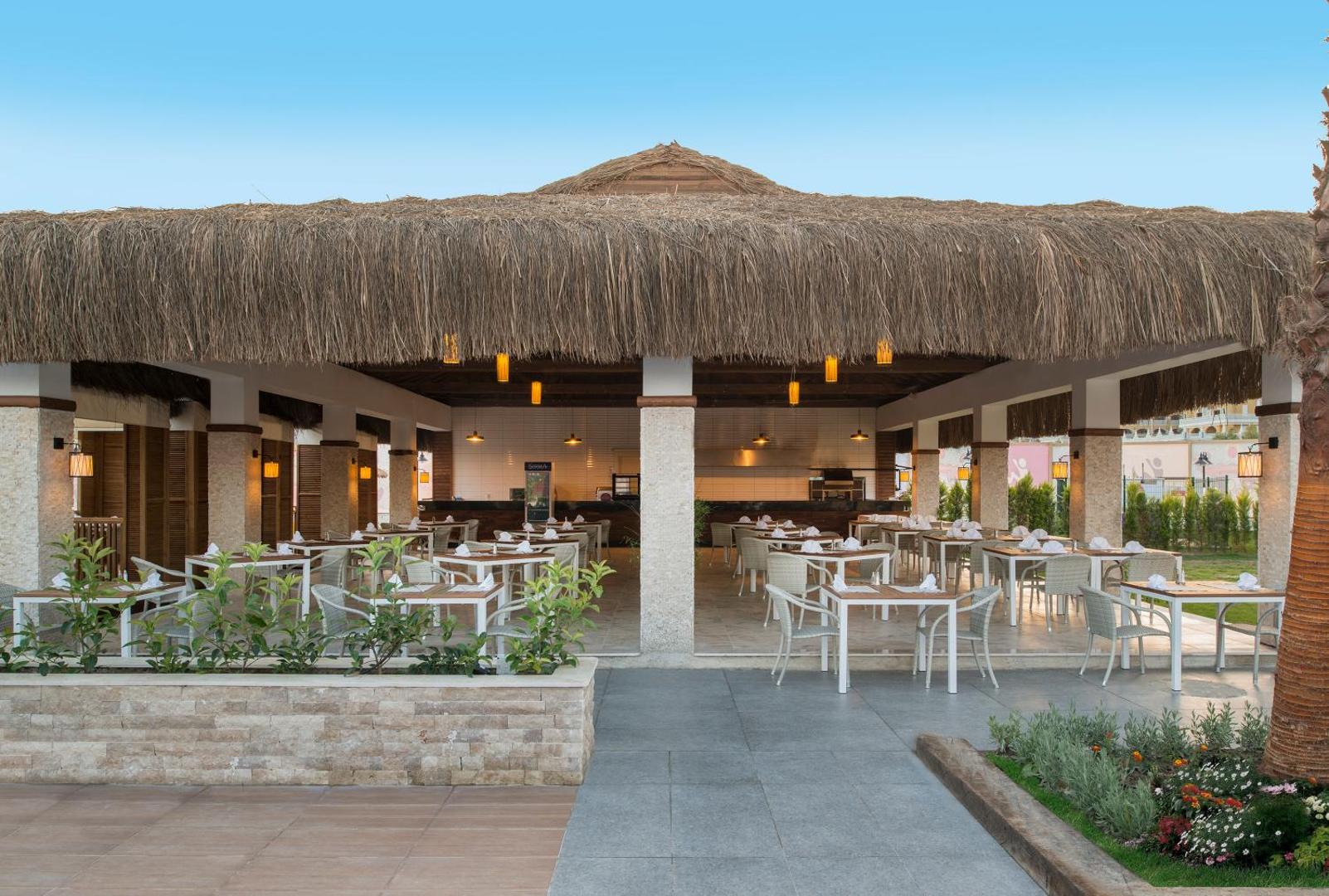 Aska Lara Resort & Spa brīvdabas restorāns ar salmu jumtu un baltiem galdiem Antālijas reģionā, Turcijā