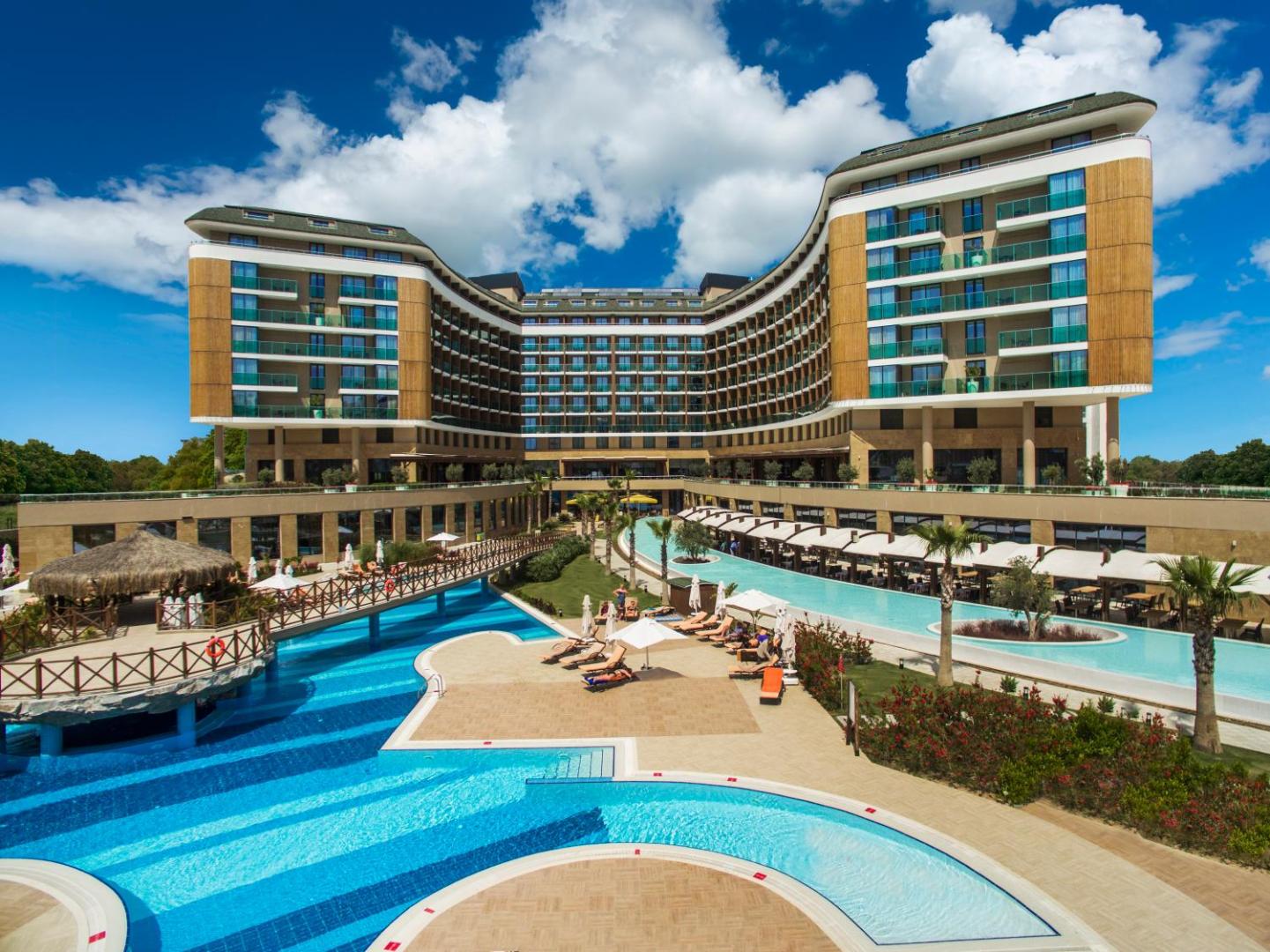 Aska Lara Resort & Spa galvenā ēka un plašais āra baseinu komplekss dienā — sauļošanās krēsli, saulessargi un palmas Turcijas piekrastē