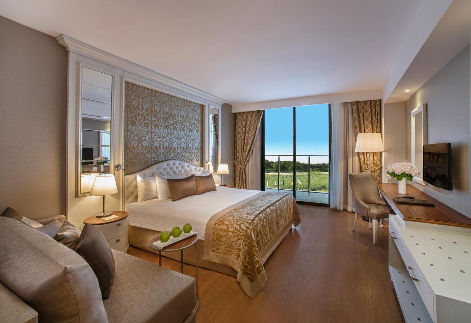 Aska Lara Resort & Spa elegants numurs ar divguļamo gultu, dīvānu un panorāmas logu uz Antālijas dabas ainavu.
