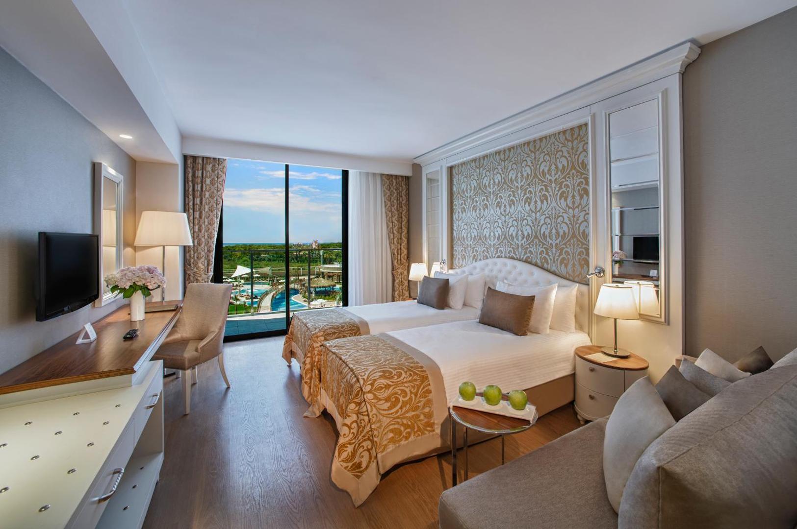 Aska Lara Resort & Spa gaišs divvietīgs numurs ar balkonu, skatu uz baseinu kompleksu un dārzu Antālijas piekrastē, Turcijā.