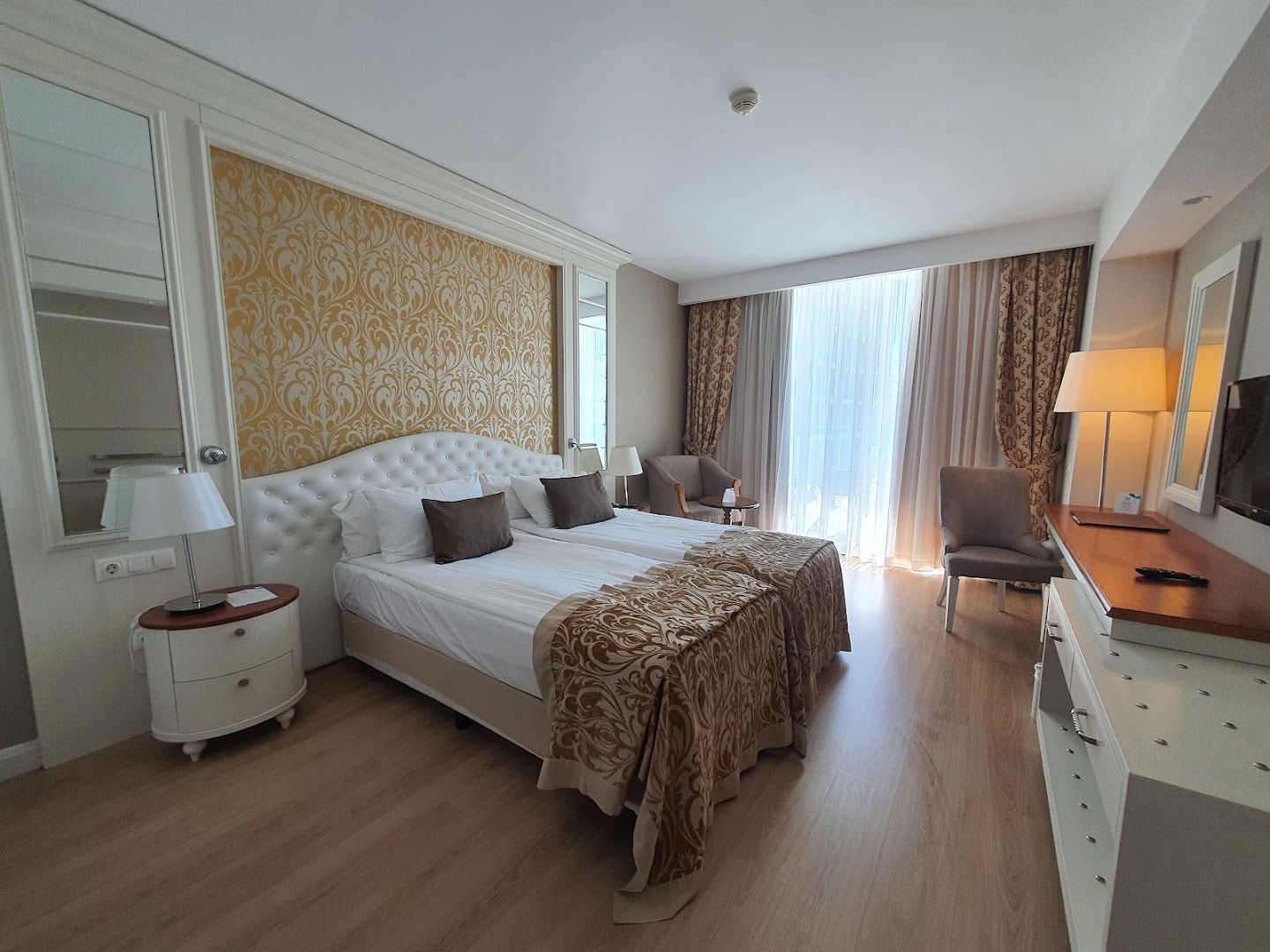Aska Lara Resort & Spa mājīgs numurs ar divām gultām, dekoratīvu sienas paneli un gaišiem aizkariem Antālijā.