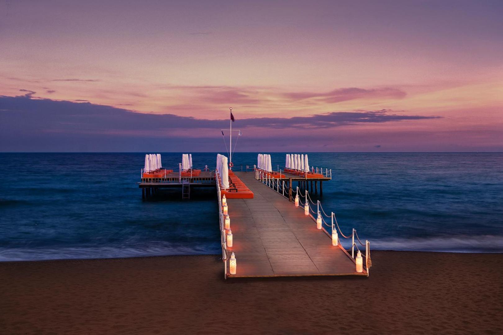 Aska Lara Resort & Spa koka mols ar sauļošanās krēsliem un laternām Vidusjūras krastā saulrieta laikā