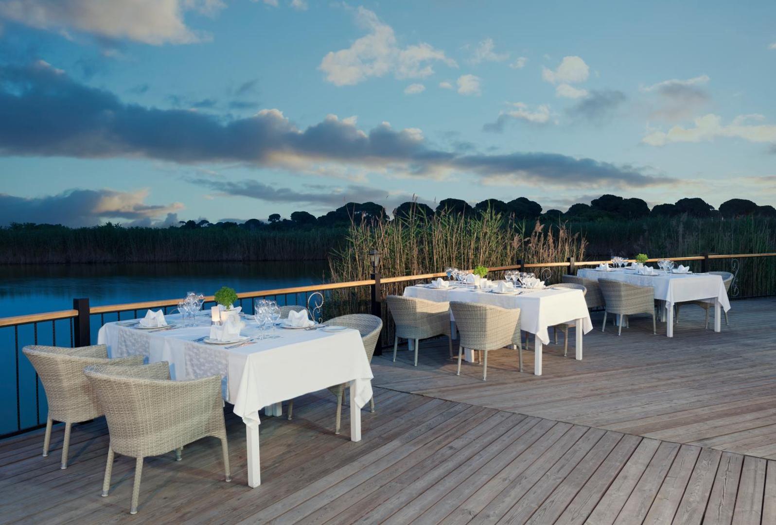 Aska Lara Resort & Spa eleganta āra terase ar klātiem galdiem pie upes vai lagūnas saulrietā