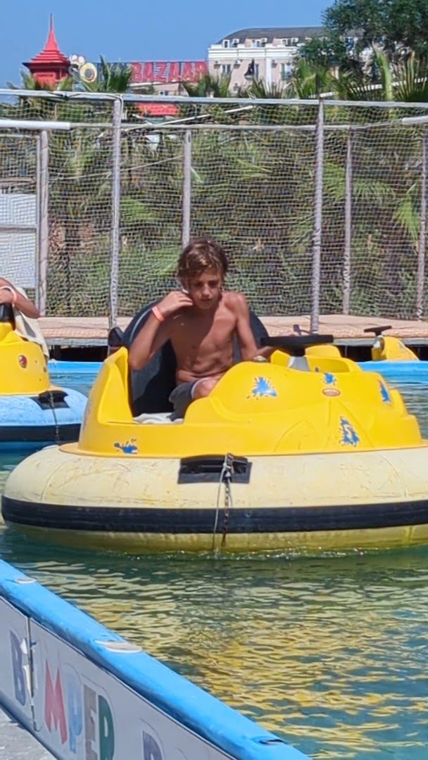 Club Aqua Plaza bērnu izklaides zona ar dzeltenām ūdens mašīnītēm baseinā, kur zēns vada vienu no tām saulainā dienā.