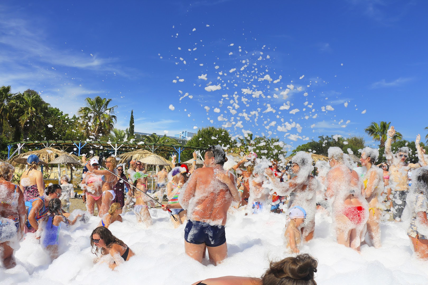 Club Aqua Plaza pludmales putu ballīte ar daudziem atpūtniekiem, kuri izbauda jautrību zem zilām debesīm un saulessargu fonā.
