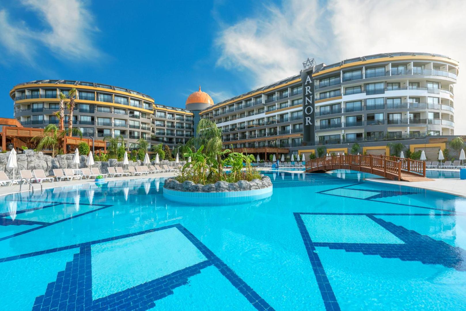Arnor De Luxe Hotel & Spa galvenais baseins ar koka tiltiņu un palmu saliņu vidū — skats uz moderno viesnīcas ēku