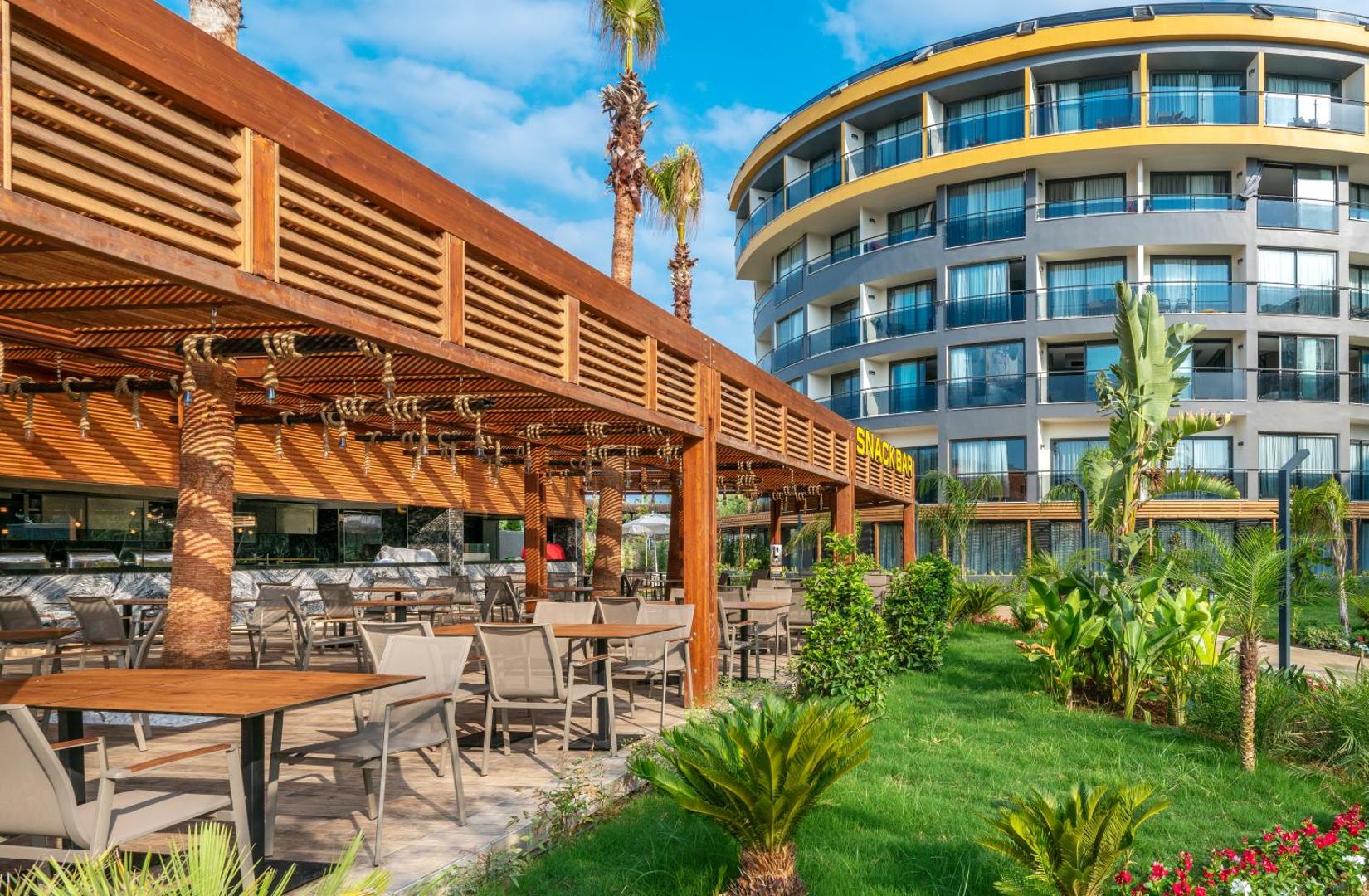 Arnor De Luxe Hotel & Spa āra uzkodu bārs ar koka nojumi, galdiņiem un palmu dārzu pie modernās viesnīcas ēkas Sidē.