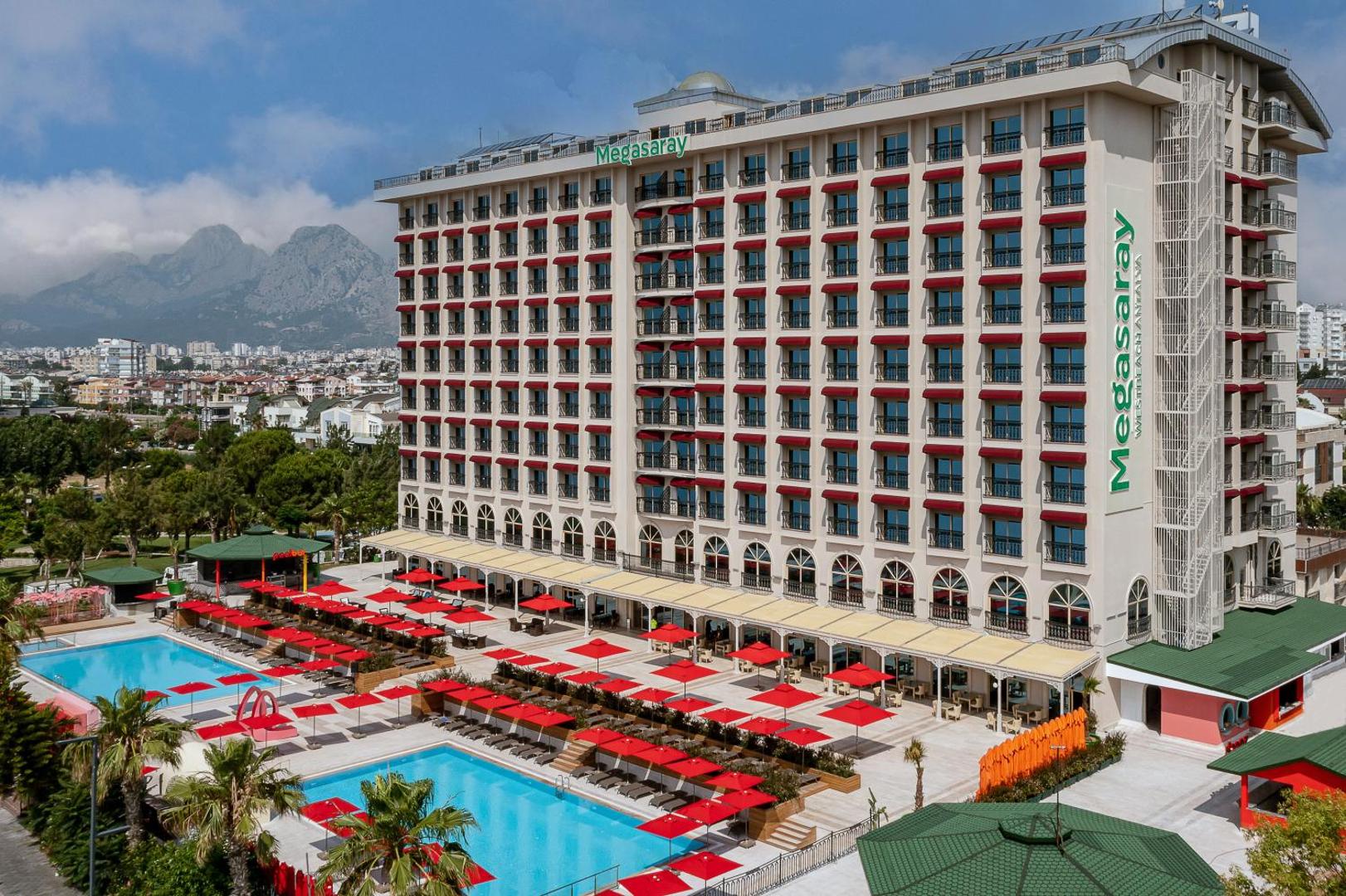 Megasaray Westbeach Antalya fasāde un atpūtas zona pie baseina ar daudziem sauļošanās krēsliem un sarkaniem saulessargiem dienas laikā.