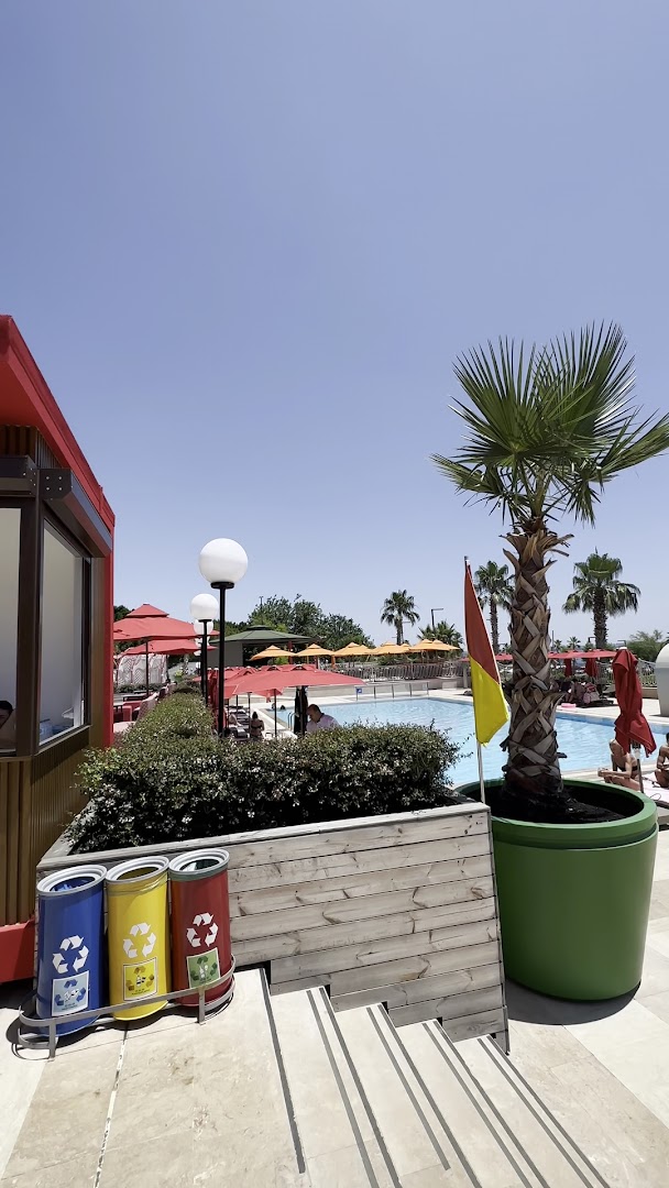 Megasaray Westbeach Antalya āra teritorija ar palmu, baseinu fonā un krāsainām atkritumu šķirošanas tvertnēm tīras vides uzturēšanai