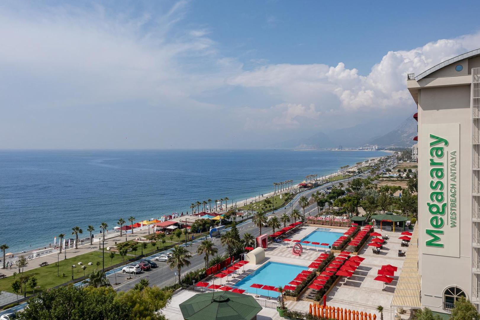 Panorāmas skats no Megasaray Westbeach Antalya terases uz pludmali, jūru un baseinu zonu ar sarkaniem akcentiem.