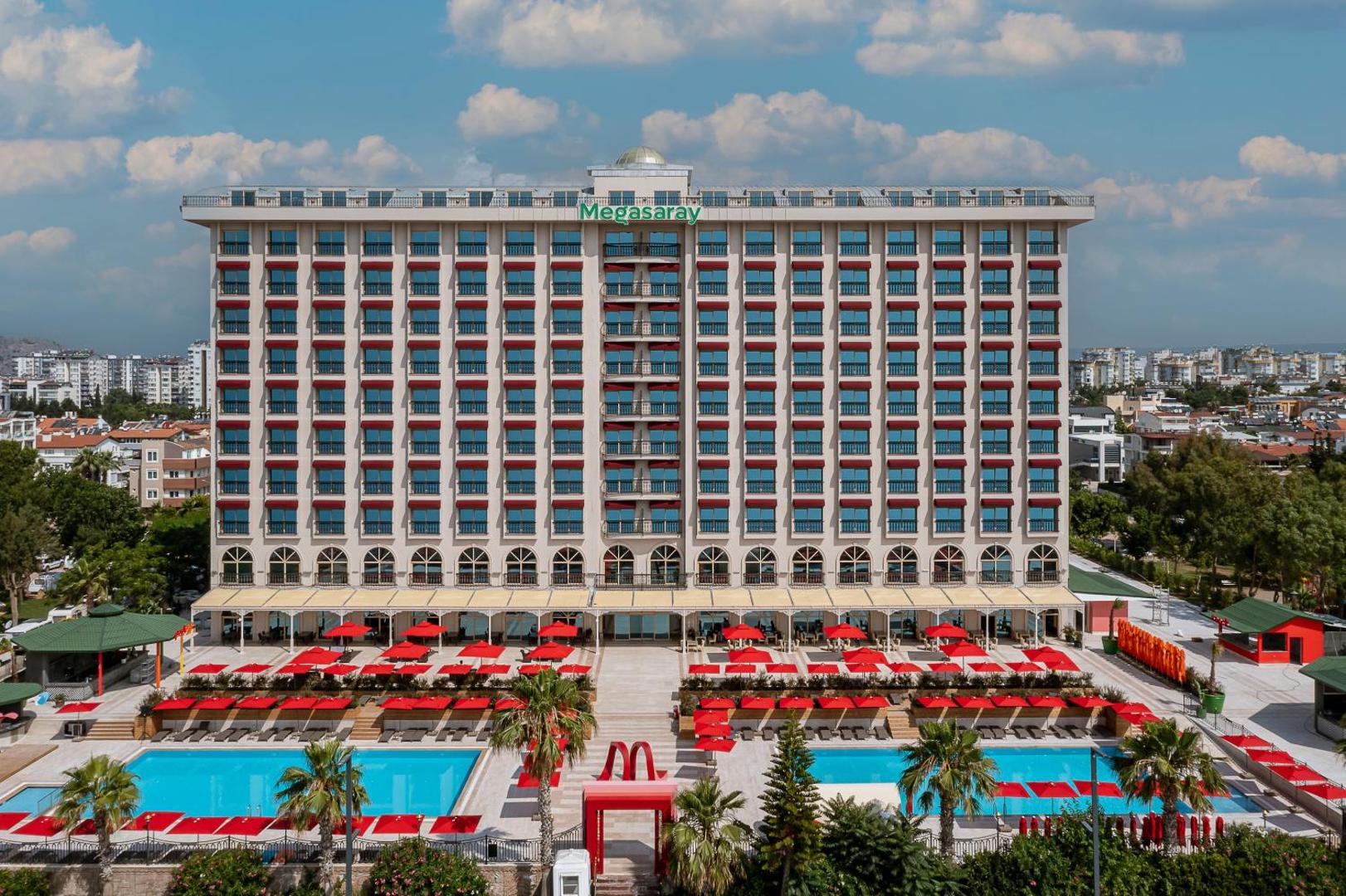 Megasaray Westbeach Antalya viesnīcas galvenā fasāde ar simetriskiem balkoniem un baseinu zonu priekšplānā zem zilām debesīm.