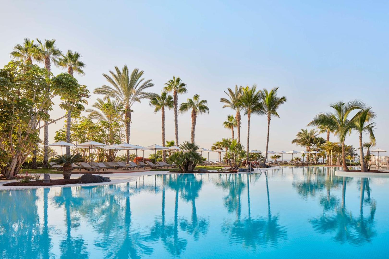 Tivoli La Caleta Tenerife Resort plašais āra baseins ar palmām un saulessargiem Tenerifes piekrastē — dzidrs ūdens un relaksējoša atpūtas zona