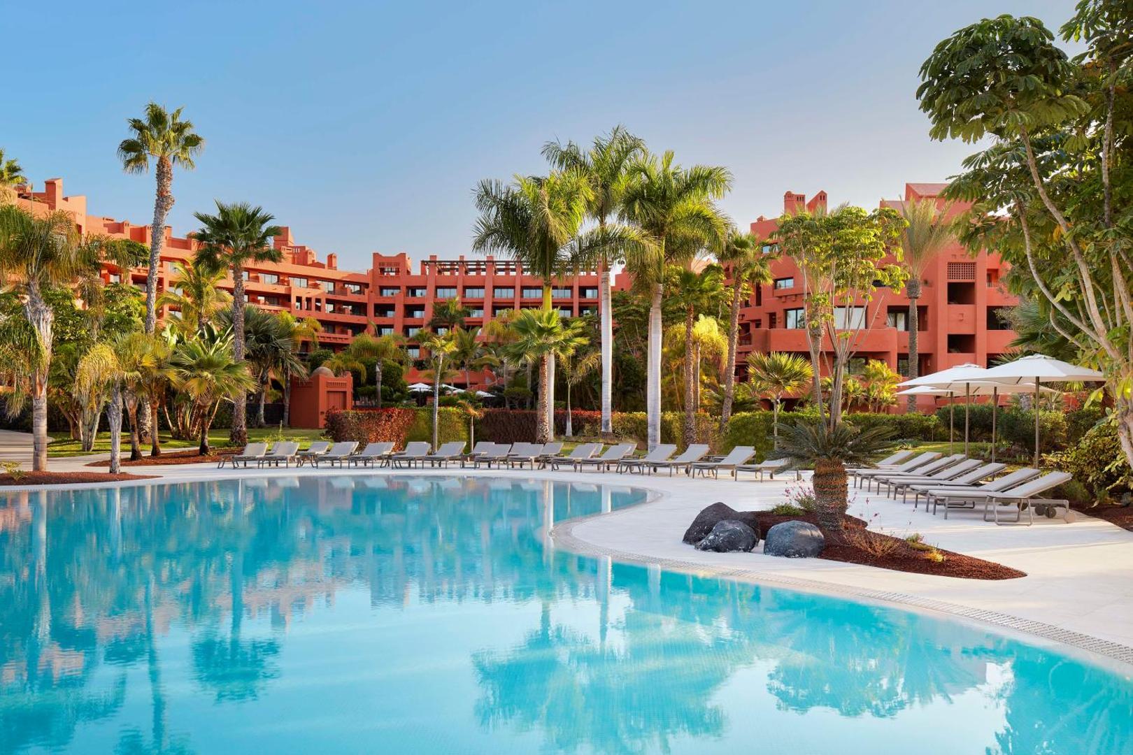 Skats uz Tivoli La Caleta Tenerife Resort terakotas krāsas ēku no baseina puses — eksotiski augi un sakopta teritorija