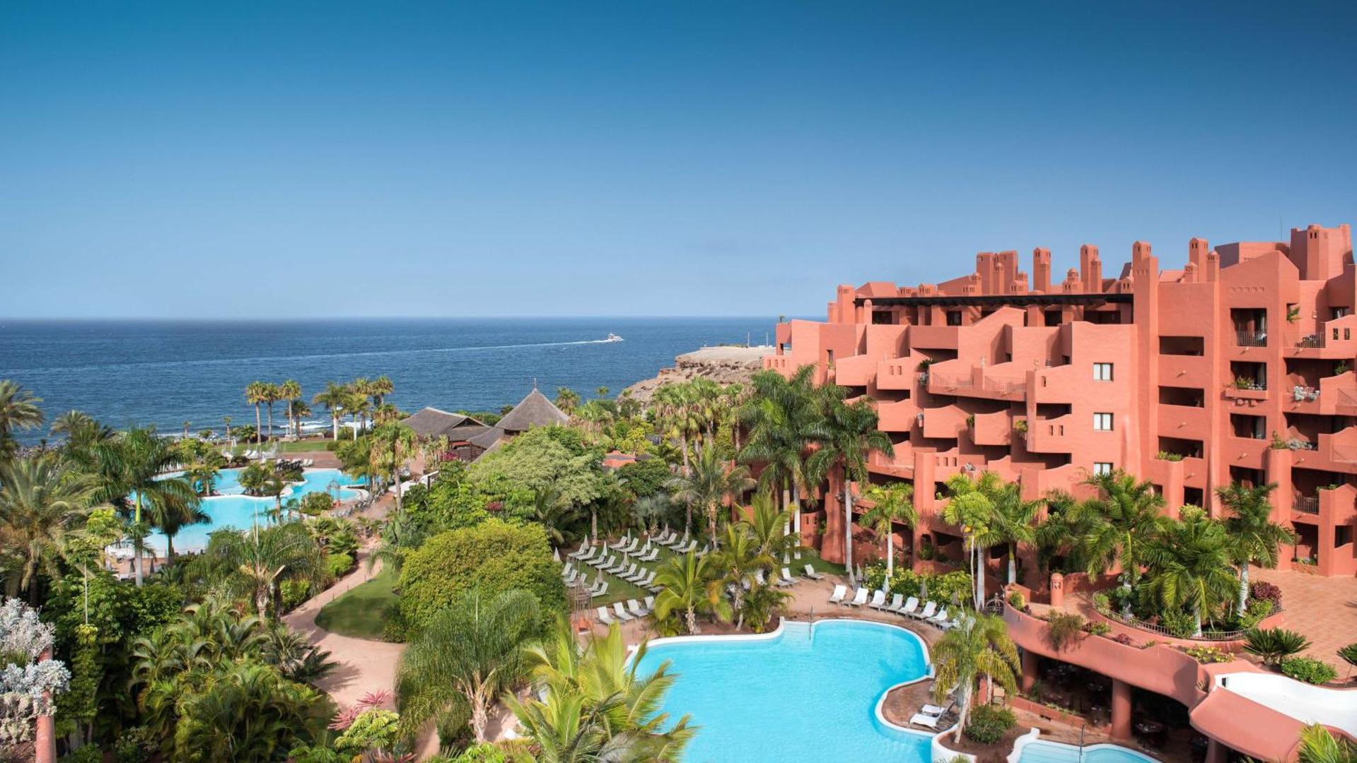 Tivoli La Caleta Tenerife Resort panorāmas skats uz baseiniem, tropisko dārzu un sārto viesnīcas korpusu pie okeāna Tenerifē.