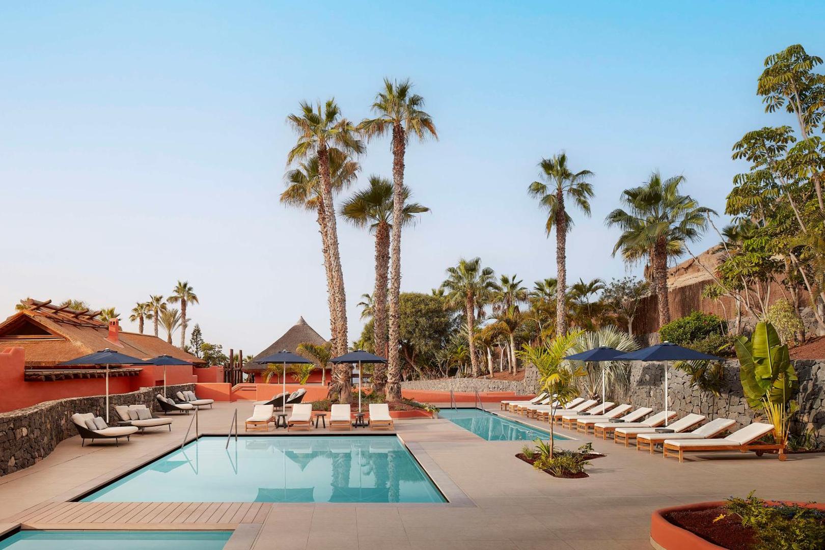 Tivoli La Caleta Tenerife Resort baseinu zona ar sauļošanās krēsliem un tumši ziliem saulessargiem — moderna arhitektūra un tropu dārzs