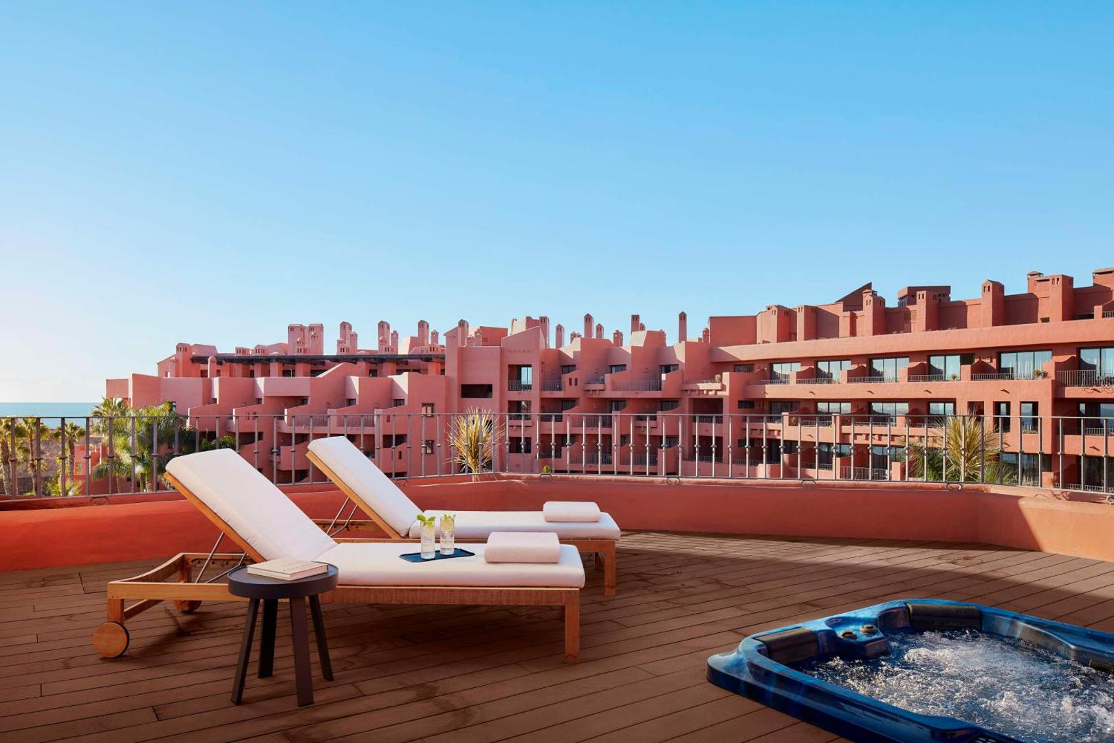 Tivoli La Caleta Tenerife Resort terase ar koka klāju, diviem sauļošanās krēsliem un burbuļvannu uz sārtas arhitektūras fona Tenerifē.