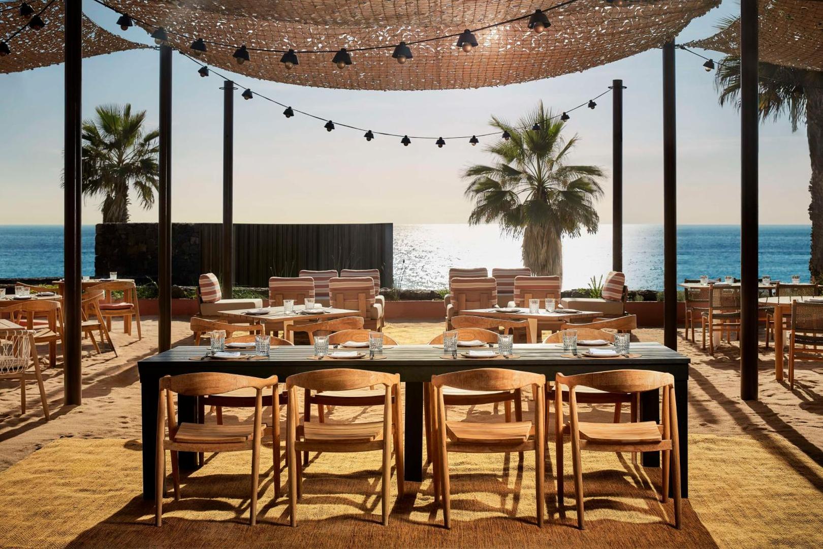 Tivoli La Caleta Tenerife Resort āra terase pludmales smiltīs ar koka galdiem un skatu uz okeānu saulrietā