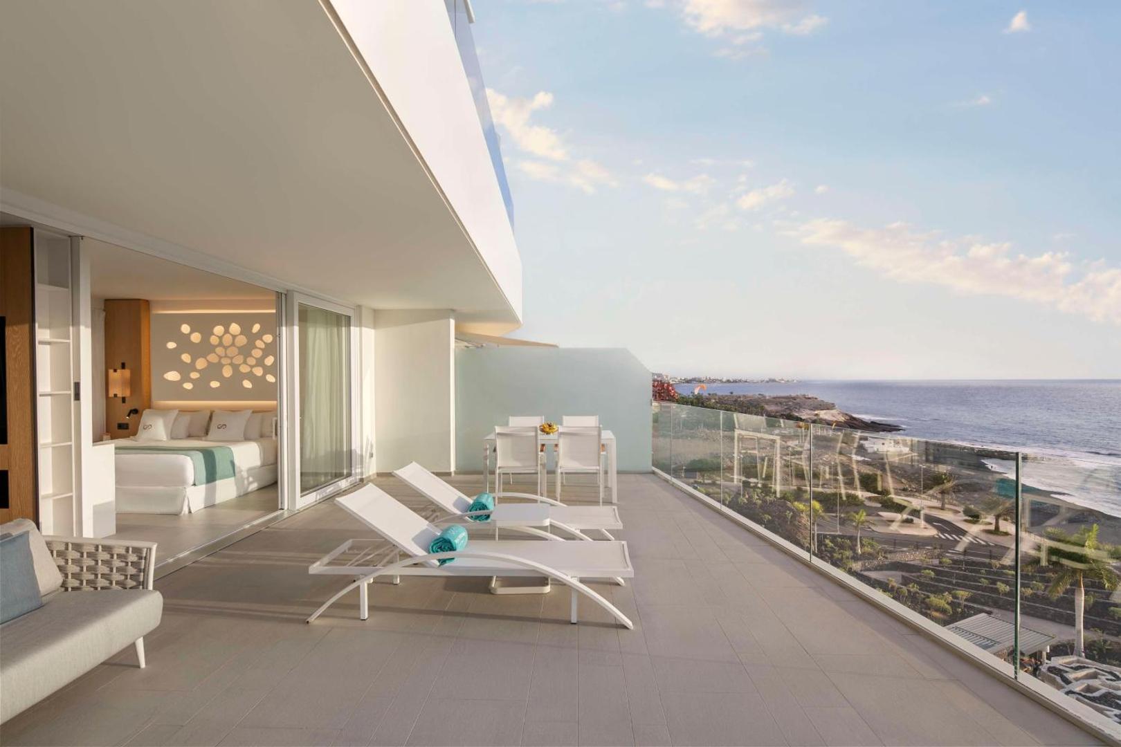 Royal Hideaway Corales Suites numura terase ar sauļošanās krēsliem un skatu uz okeānu Tenerifē — gaišs dizains un stikla margas