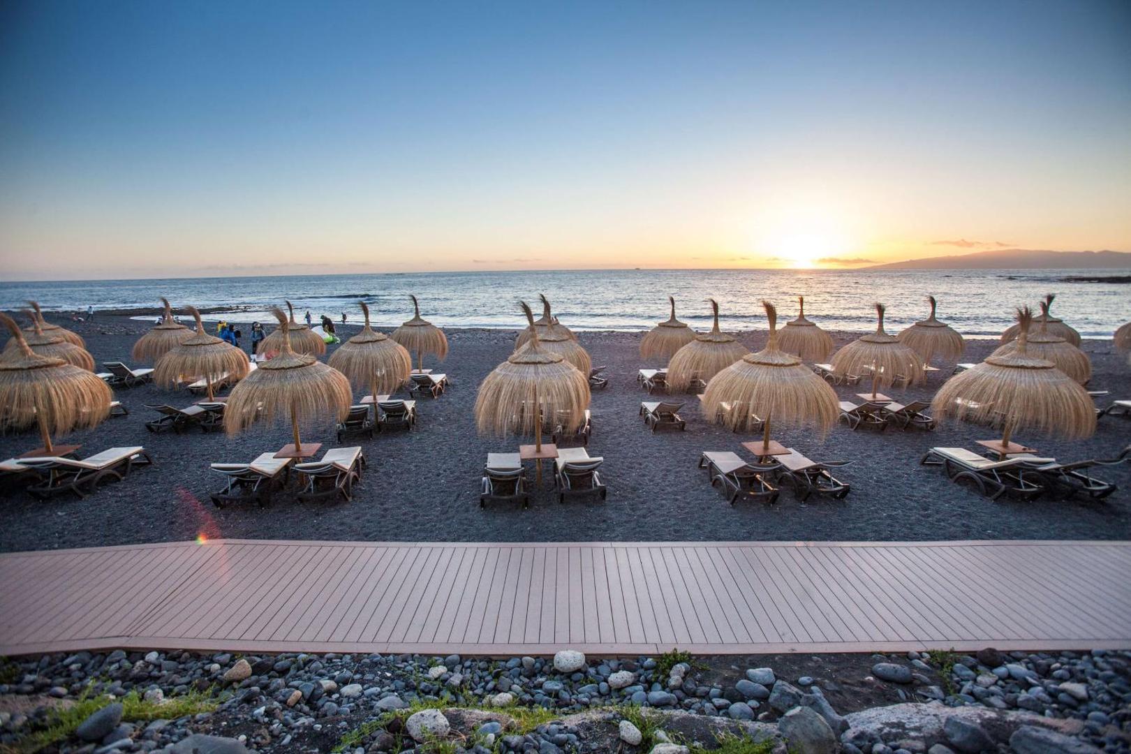 Royal Hideaway Corales Beach pludmales zona saulrieta laikā Tenerifē — salmu saulessargi un zviļņi uz tumšo smilšu krasta pie okeāna
