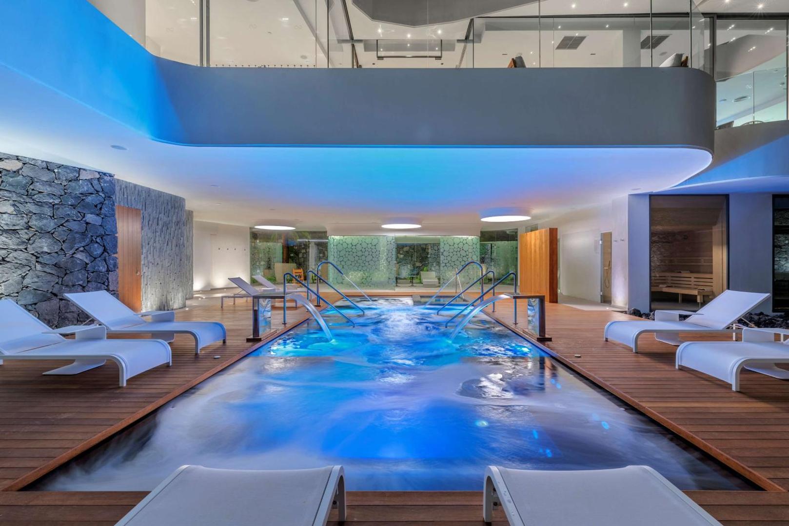 Royal Hideaway Corales Beach spa centrs Tenerifē ar iekštelpu hidromasāžas baseinu, atpūtas krēsliem un zilu apgaismojumu relaksācijai.