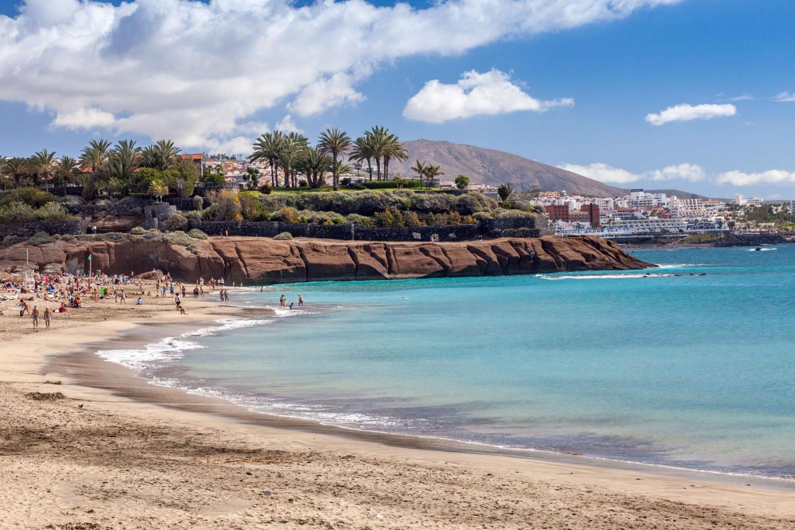 Skats uz publisko smilšu pludmali un Royal Hideaway Corales Beach apkārtni Tenerifē — tirkīza ūdens, klintis un palmas piekrastē