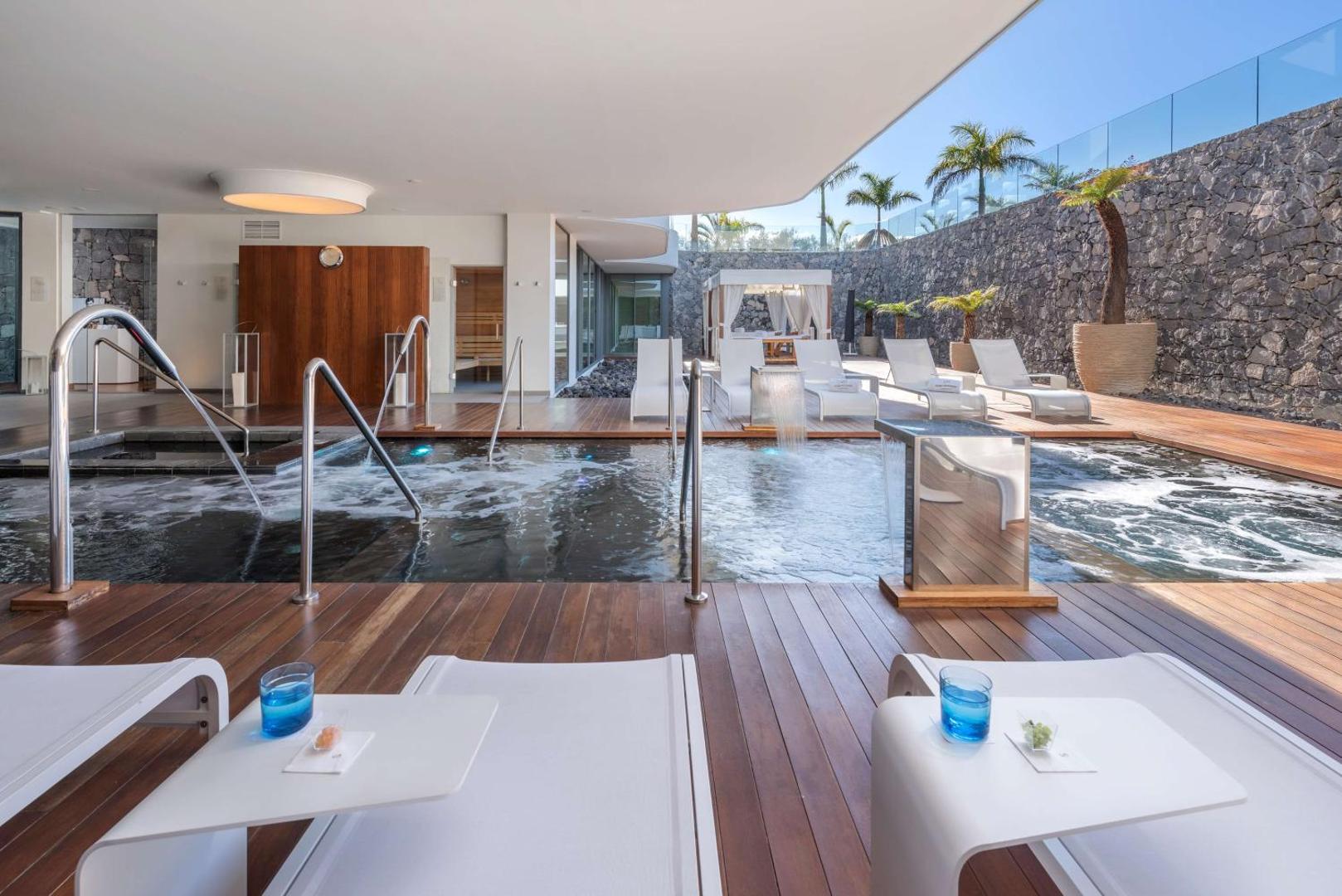 Royal Hideaway Corales Beach āra spa zona ar baseinu, koka terasi, sauļošanās krēsliem un akmens sienas dekoru Tenerifē.