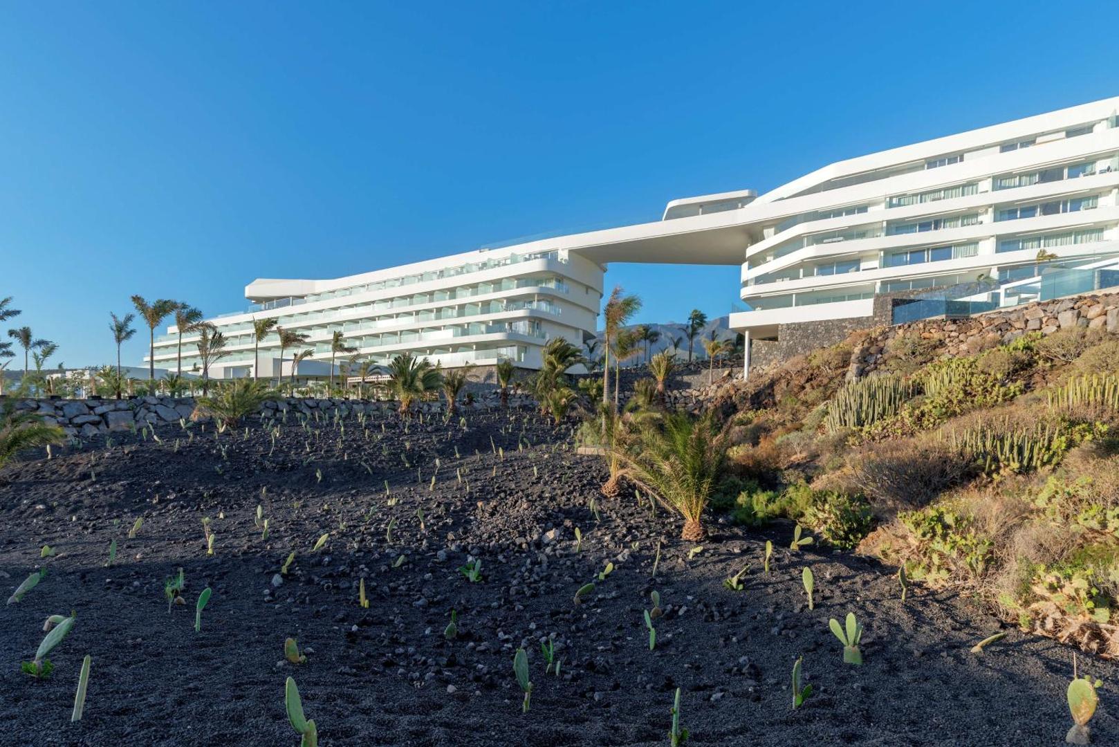 Royal Hideaway Corales Beach viesnīcas ēka dienas laikā — balta, viļņveida arhitektūra uz vulkānisko iežu un palmu fona