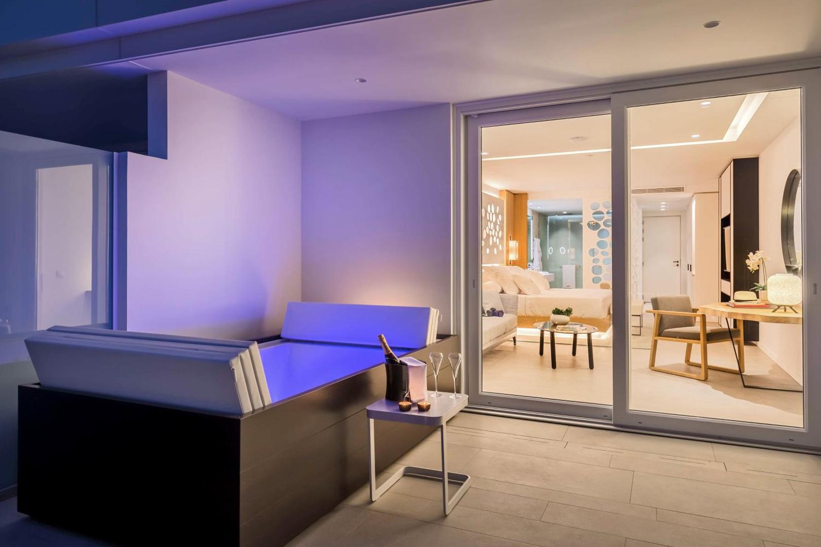Royal Hideaway Corales Beach numura terase vakarā ar izgaismotu āra vannu, šampanieti un skatu uz izgaismoto numura interjeru