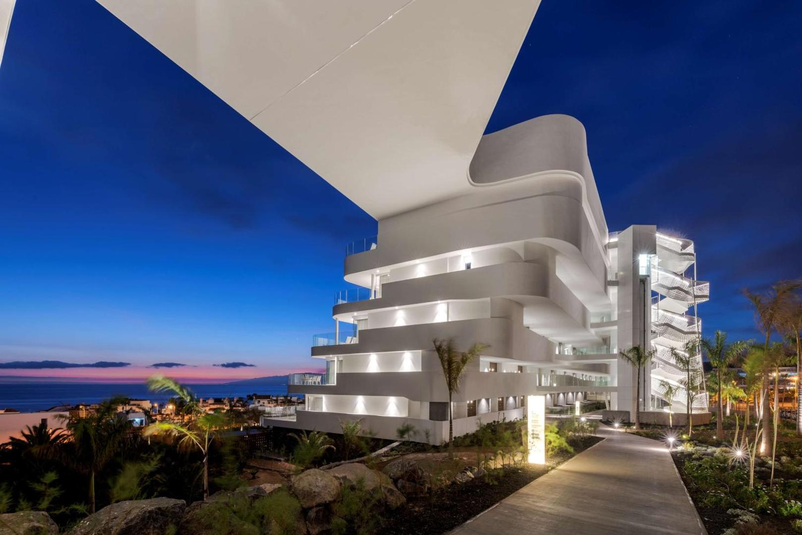 Royal Hideaway Corales Beach modernā arhitektūra vakarā Tenerifē — izgaismotas terases un numuri ar skatu uz okeānu saulrieta laikā
