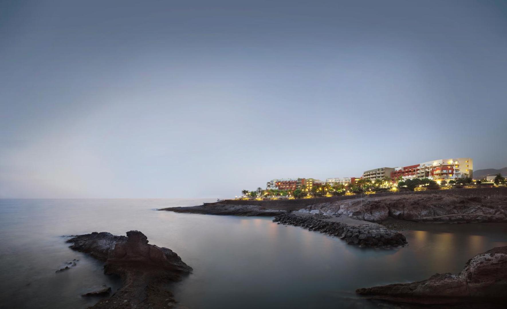 Viesnīcas Roca Nivaria panorāmas skats krēslas stundā Tenerifes piekrastē — izgaismota ēka pie okeāna līča ar akmeņainu krastu
