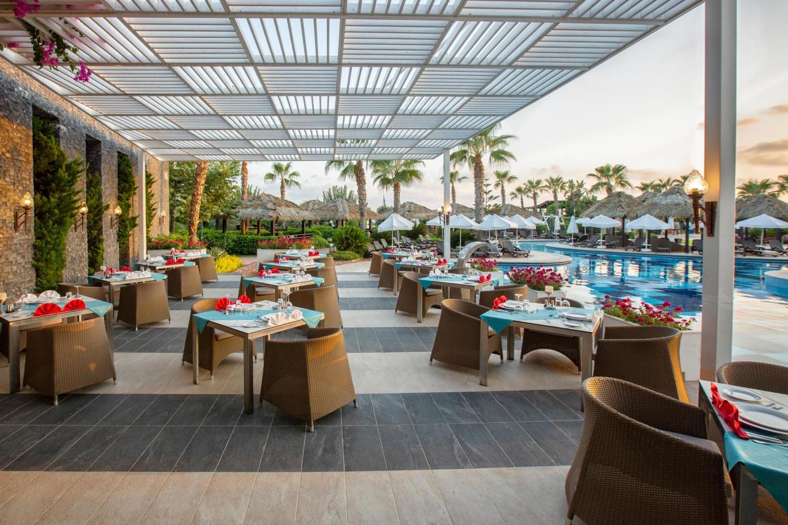 Sherwood Blue Belek āra restorāna terase ar skatu uz baseinu un palmām saulrietā — eleganti klāti galdi zem nojumes
