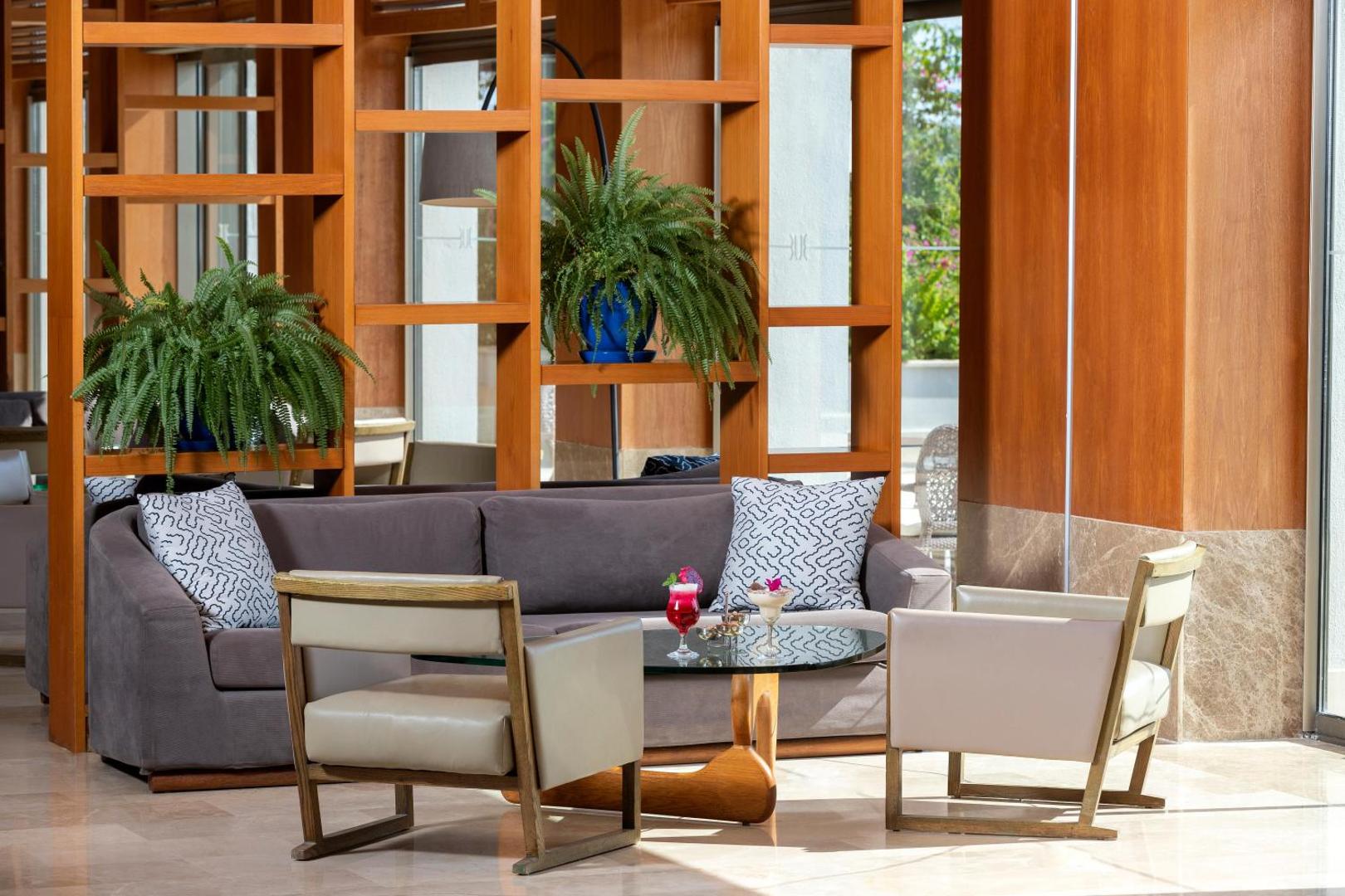 Sherwood Blue Belek viesnīcas lobby atpūtas zona ar mīkstajiem dīvāniem, krēsliem un dekoratīviem telpaugiem koka plauktos Belekā, Turcijā