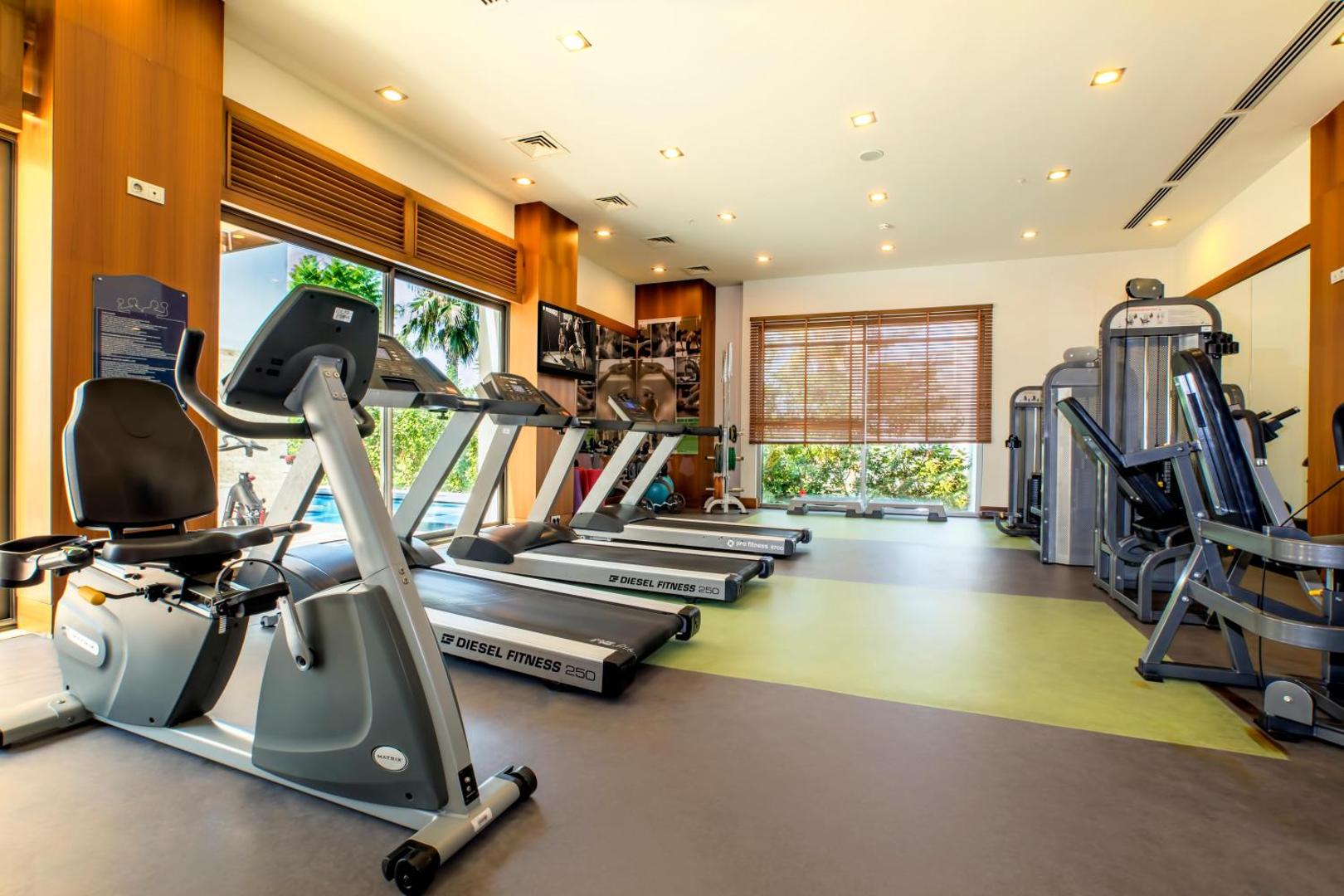 Sherwood Blue Belek fitness centrs ar moderniem skrejceliņiem un trenažieriem gaišā telpā ar skatu uz dārzu