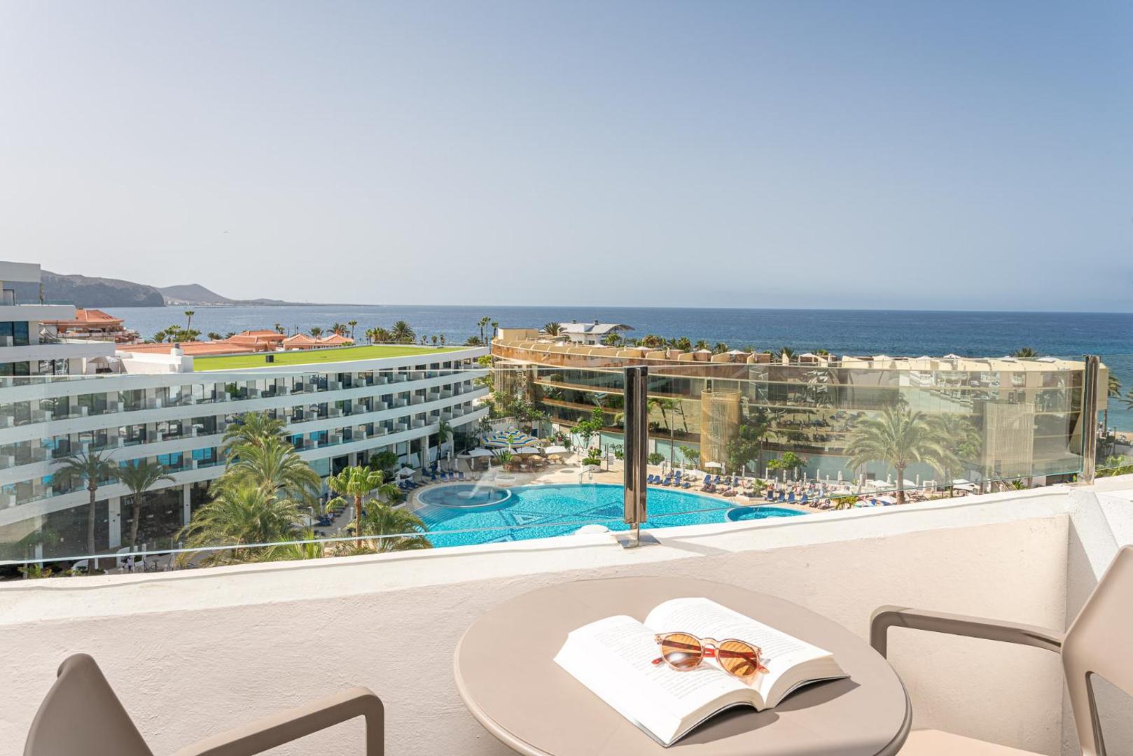 Skats no Mediterranean Palace balkona uz viesnīcas baseinu kompleksu un okeānu Tenerifē — atpūtas zona ar galdiņu un grāmatu