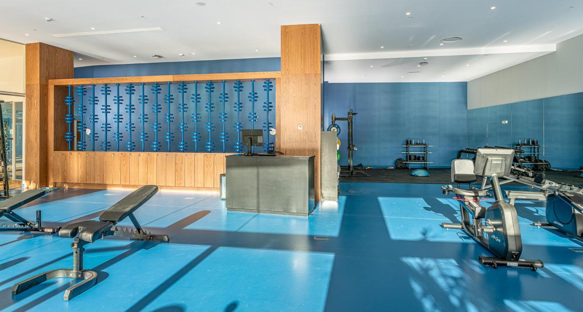 Plašs Mediterranean Palace viesnīcas fitness centrs ar zilu grīdas segumu, svaru stieņiem un trenažieriem spēka treniņiem