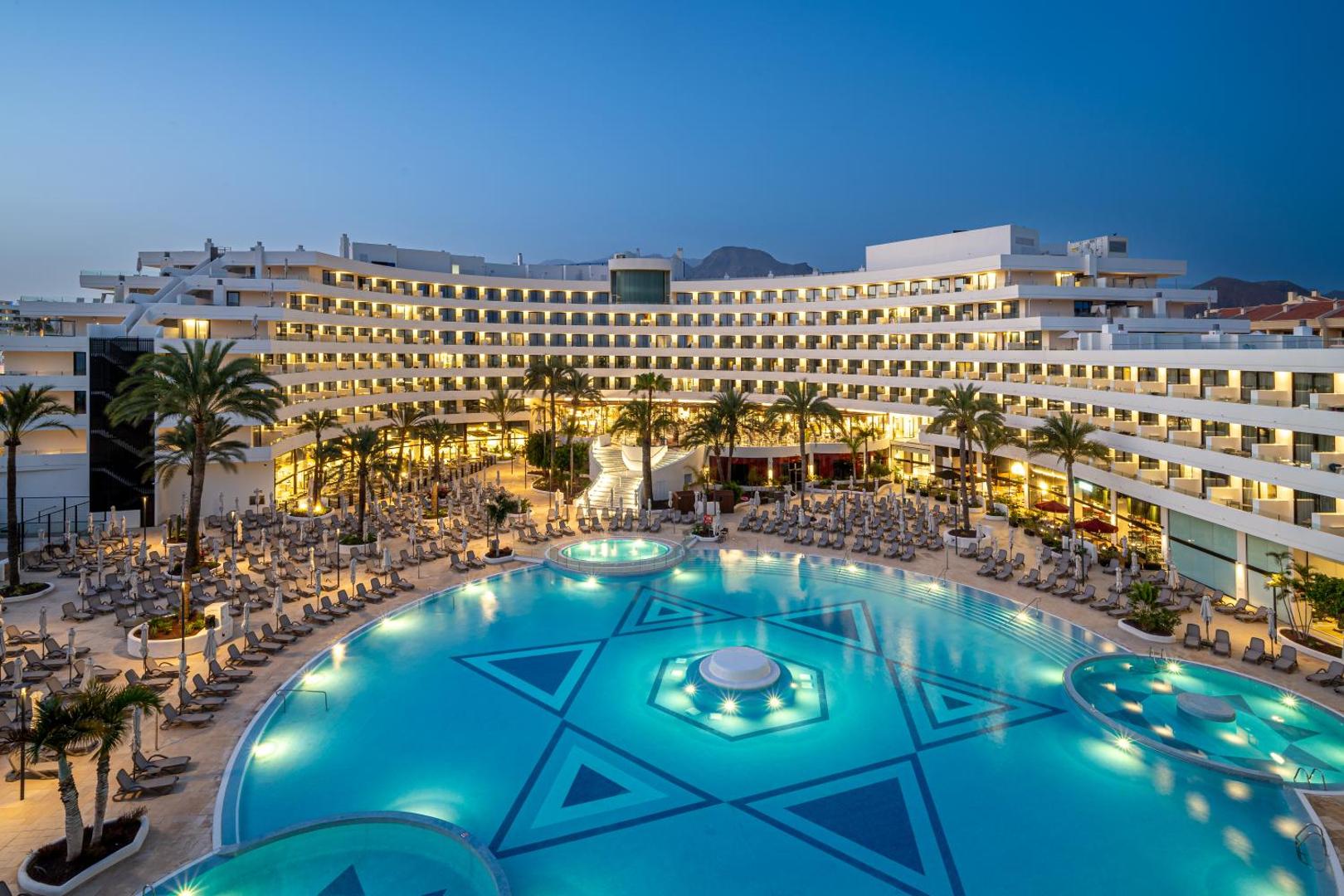 Mediterranean Palace viesnīcas plašais āra baseins un izgaismotā fasāde vakarā Tenerifē, Spānijā — redzami sauļošanās krēsli un palmas.