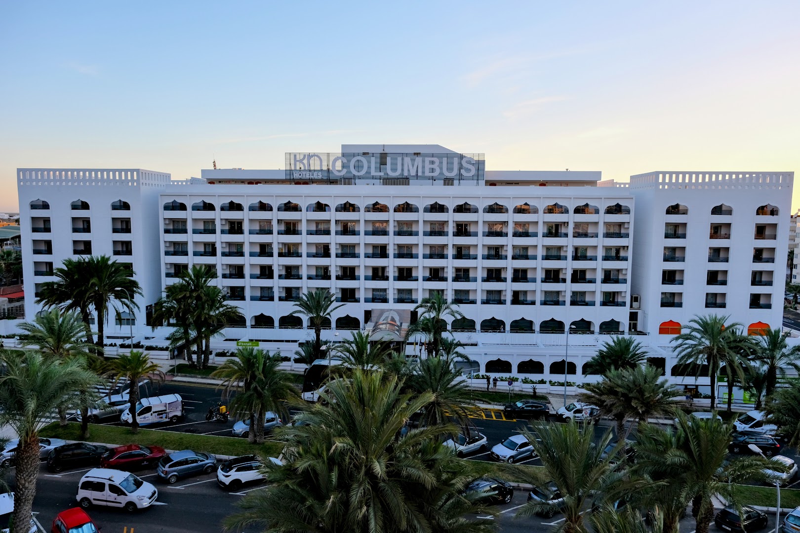 Kn Columbus Aparthotel ēkas fasāde no attāluma Tenerifē — balta arhitektūra ar arkveida balkoniem, palmām un automašīnu stāvvietu priekšplānā