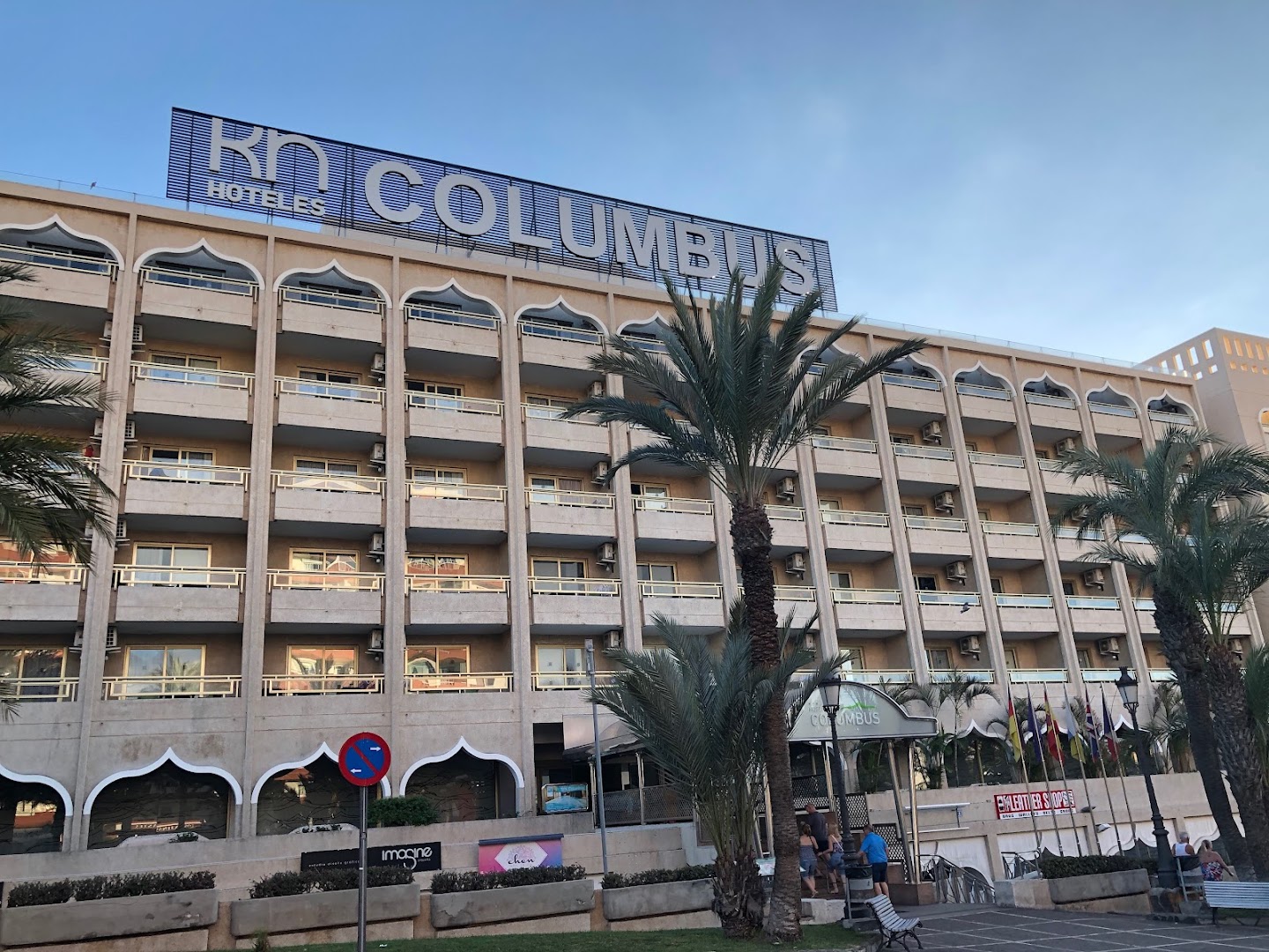 Kn Columbus Aparthotel daudzstāvu ēka ar balkoniem un palmām vakara gaismā — redzama viesnīcas izkārtne un apkārtējā infrastruktūra