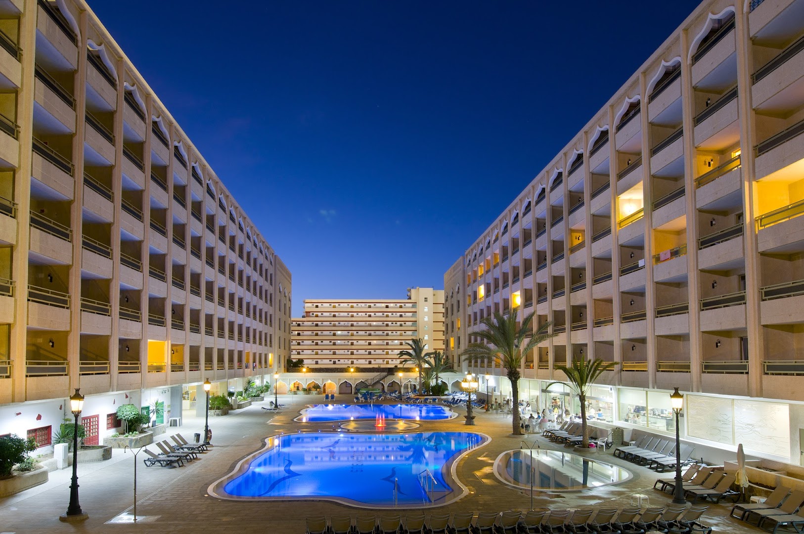 Kn Columbus Aparthotel iekšpagalms vakarā ar izgaismotiem baseiniem un viesnīcas korpusiem Playa de las Americas kūrortā Tenerifē