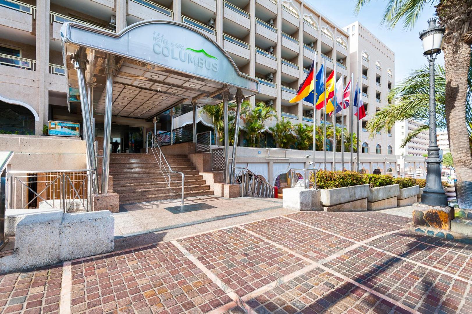 Viesnīcas Kn Columbus Aparthotel galvenā ieeja Tenerifē ar kāpnēm, metāla nojumi, karogiem un palmām saulainā dienā