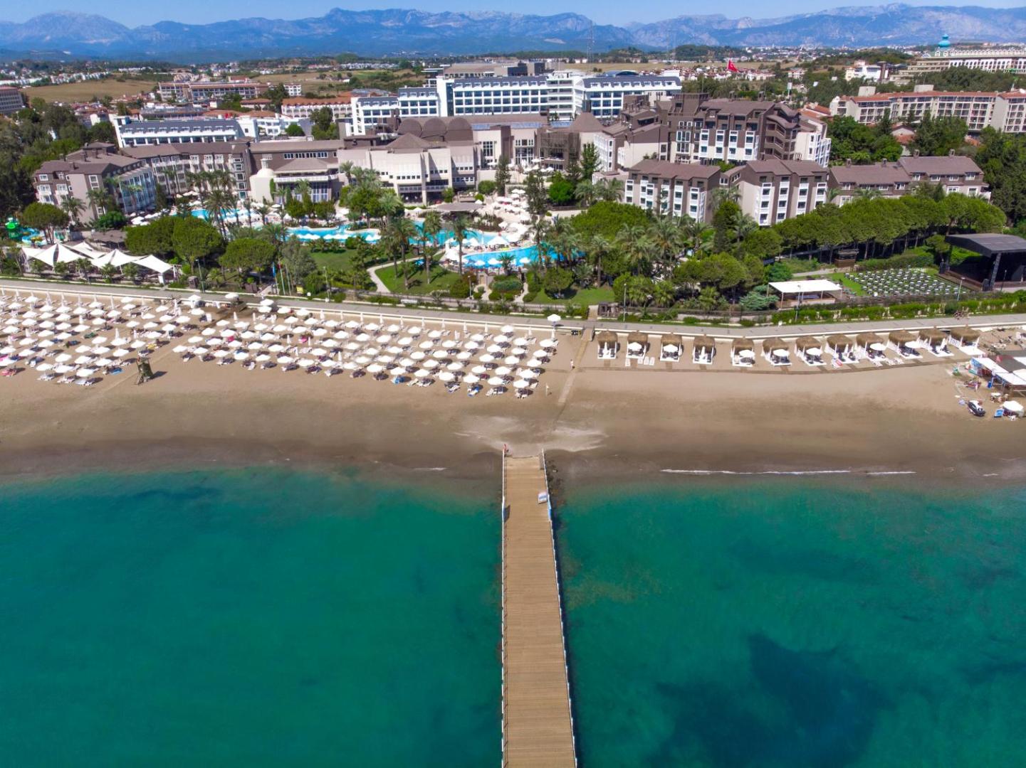 Paloma Perissia viesnīcas panorāmas skats Side, Turcijā — plaša smilšu pludmale ar saulessargiem, koka piestātne un baseinu komplekss pie jūras