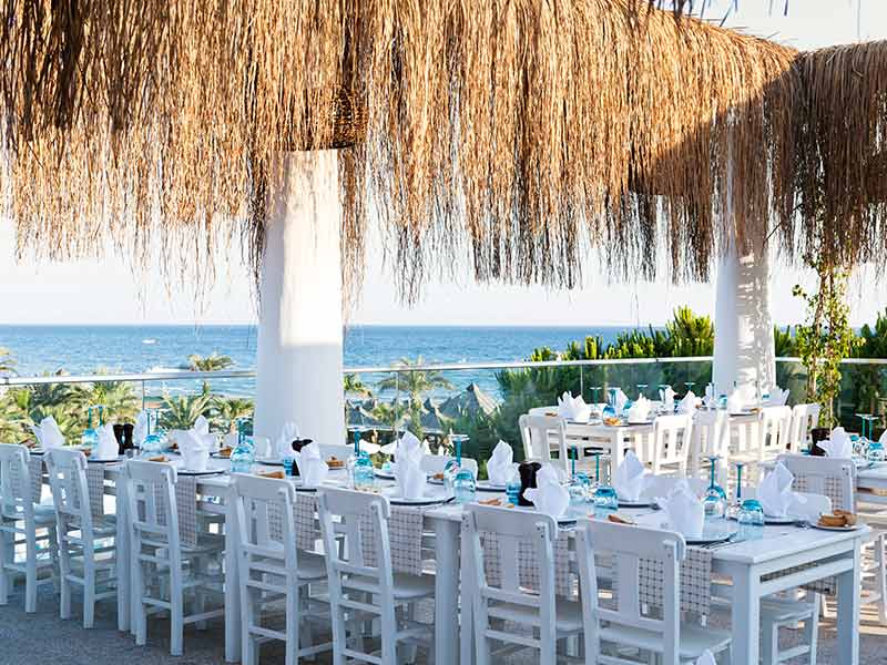 Paloma Oceana brīvdabas restorāna terase ar skatu uz jūru, balti klātiem galdiem un tradicionālu niedru jumtu