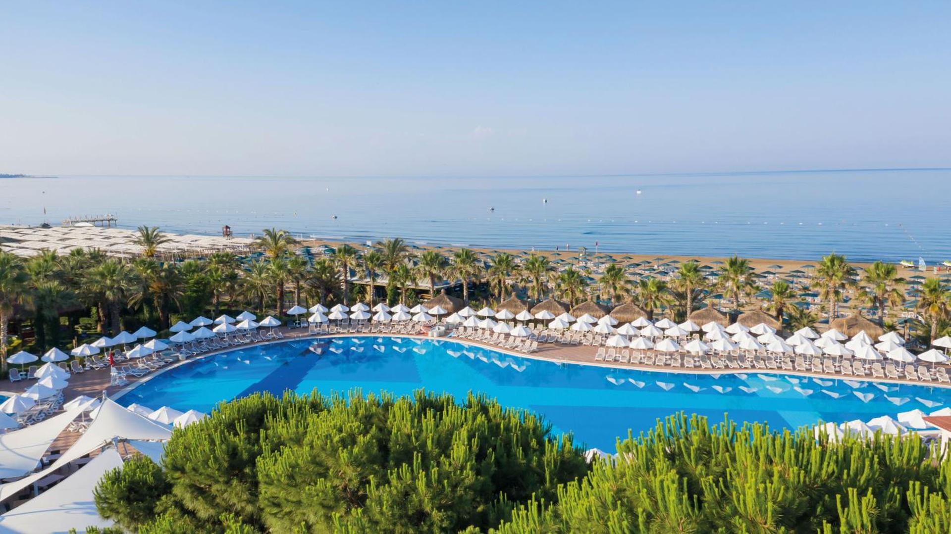 Paloma Oceana viesnīcas panorāmas skats uz lielo āra baseinu, smilšu pludmali un Vidusjūru Sidē, Turcijā — apkārt palmas un saulessargi